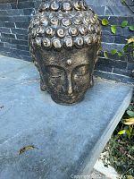 Buddha head statue sitting on a concrete ledge against a dark stone wall with some greenery in background.