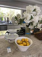 Image of two white ceramic cake stands stacked, large white bowl with lemons inside, clear glass vase with faux white flowers on kitchen counter.