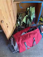 Three folded green folding camp chairs leaning against a wooden wall with a large red and gray carrying bag on the floor.