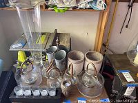 Photo showing all items in the lot placed on a wooden surface and shelf behind. Includes two large glass lanterns, two ceramic candle holders, two metal buckets, the Buddha head candle holder, and a small mercury glass votive candle.