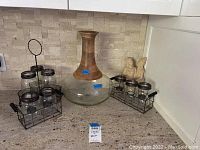 Photo showing two sets of three clear glass jars each inside black metal wire holders with side handles, containing white candles, positioned on kitchen counter next to large glass vase and angel decoration.