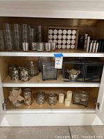 Three shelves in a wooden cabinet containing various candles and candle holders including a box of white tealights, glass holders, pillar candles, and metal holders.
