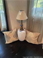 White ceramic textured table base with attached brown lamp base and neutral tone lampshade. Two beige pillows with embroidery placed on floor beside table.