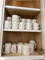 Shelf with 14 plain white ceramic coffee mugs arranged in two rows, some stacked.