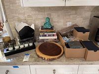 Kitchen counter with cookbooks stacked beside a turquoise Buddha statue, a wooden round board, and small glass candle holders on a black stand, with cardboard boxes containing other items.