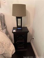 View of dark wood end table and associated modern lamp on top, positioned next to bed and wall.