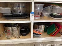 Clear and white plastic food storage containers, black metal baking trays, glass measuring cup, metal grater, small food chopper, metal pots with colored lids on a kitchen shelf.