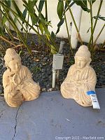 Two seated Buddha statues on a stone surface surrounded by stones and plants, showing overall wear and scratches.