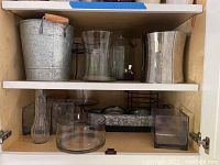 Shelf displaying galvanized metal ice bucket with wooden handle, clear glass ice bucket with spigot, clear glass candle holders, black metal wire rack, metal tray, and square glass container.