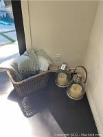 Basket with two light-colored throws beside two glass and metal lanterns with candles inside, placed on dark floor near a wall.