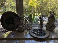 Full view of lot items: painted plaque, two metal lanterns, decorative grate, vase with yellow dried flowers, white owl sculpture on marble surface near window