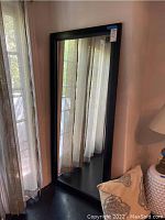 Full front view of tall rectangular leaning wall mirror with black frame next to window with light curtains.