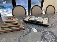 Set of two mirrored trays and stack of ten assorted picture frames on table with chairs and glass door background.