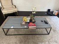 Rustic coffee table with gray wooden plank top holding decorative items such as flowers, candles, and books on a patterned rug with a beige upholstered chair in the background.