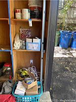 Lower wooden cabinet shelf and floor area with Ryobi power saw, cords, light bulbs, boxes, bags