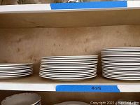 Photo of stacks of white dinner plates, salad plates, and bowls arranged on wooden shelves, showing three groups of plates and one group of bowls.