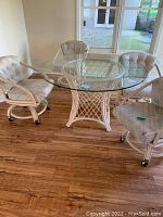 Full view of round glass top dining table with wooden lattice base and four matching cushioned swivel chairs with casters inside a room with wood floor and windows.