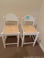 Two white wooden barstools with woven seats placed side by side against a beige wall and carpeted floor. Both show spots on the woven seat material.