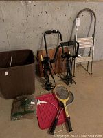 Photo showing metal step stool, two bike racks, two sports racquets, pink snow shovel, large plastic tub, plastic bag with greenery, and a white handheld device arranged on concrete floor against cement wall