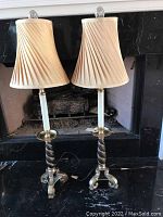 Pair of buffet lamps with beige pleated fabric shades and tall golden black tone twisted bases placed on dark surface in front of fireplace.