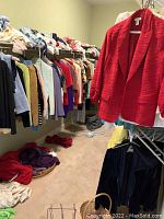 View of multiple ladies clothing items hung on racks with a wide range of colors including a prominent red jacket in the foreground and other tops and sweaters along a rack against a wall.