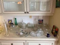 Overview of lot showing multiple crystal bowls, glass jars, acrylic carafe, pewter steins, and Pyrex dish on kitchen counter