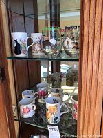 Shelf showing two cat decorated mugs, two stemless cat decorated glasses, and floral decorated mugs below.