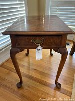 Full view of the Hekman wood end table showing polished wood grain top, cabriole legs, metal drawer pull and tag.