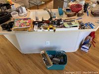 Wide view of kitchen counter with various kitchen utensils, some cutting boards, and kitchen tools displayed. Plastic bin on floor contains more cutlery and utensils.