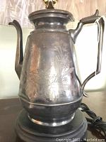 Close-up photo of the ornate engraved metal coffee pot used as the lamp base, showing the handle, spout, and decorative floral patterns.