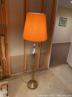 Full view of one brass floor lamp with orange pleated shade standing on concrete floor against unfinished wall.