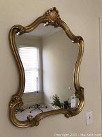 Front view of the gold plastic framed mirror hanging on wall, showing ornate curved frame details and reflection of room window.