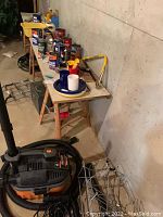 Wide shot of basement corner showing Shop Vac vacuum, table with cans and boxes, coiled hose, and metal box.