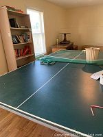 Full view of green ping-pong table with white markings, paddles, and extra net placed on top.