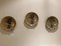 Three oriental-style decorative plates with metal wall hangers mounted on wall; two plates have pineapple plant design and one with palm tree design.