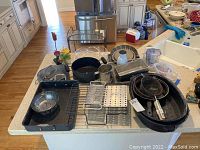 Various cookware items arranged on kitchen counter including roasting pans, strainers, graters, baking pans, measuring cup, and chipper.