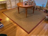 Full view of the rectangular sisal rug under a wooden round dining table in a kitchen setting, showing its natural tan color with a red-brown border and visible wear marks.