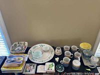 Wide view of assorted ceramic serving dishes, bowls, cups, decorative tiles and coasters displayed on a black table against beige wall.