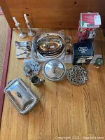 Full view of all items on wooden floor including silver plate serving dish, pewter pitcher, candle sticks, decorative trays, boxed set, and other small pewter items.