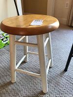 Small round wooden stool with natural wood seat and white painted legs, paint splatters on legs, positioned on carpeted floor near wall with electrical outlet.