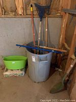 Multiple yard tools grouped around a large grey trash can including shovels and a blue rake, with green bucket and sand bag on floor.