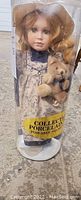 Porcelain doll in clear plastic display case showing doll with hat holding teddy bear, label reads 'Collectable Porcelain Posable Stand'