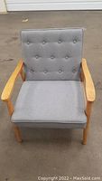 Gray cushioned chair with button-tufted backrest, wooden armrests and legs, displayed on showroom floor.