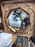 Photo of the octagon shaped mirror with a gold geometric frame shown inside a cardboard box with some packaging materials. The mirror surface reflects surroundings clearly.