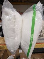 Two new soft pillows in plastic packaging side by side on shelf.