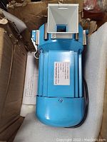 Front angled view of a blue electric soil and spice grinder showing the motor housing and feeding hopper inside an open box with protective foam.