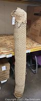 Photo shows a rolled-up brown woven rug standing upright on a shelf in a warehouse-like setting. The rug appears tightly rolled with a visible woven texture and is wrapped with some of its original packing material at the top.