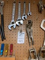 Photo shows four adjustable wrenches, multiple locking pliers, a C-clamp locking plier, and three screwdrivers with colored handles on pegboard. Small clip with lot number attached.
