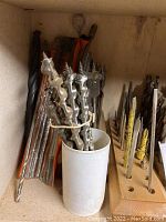 Large auger drill bits held in a white cylindrical container with additional long drill bits in the back.