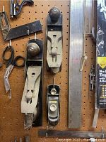 Metal hand planes, steel square, metal ruler, scissors, compass divider, and handsaw hanging on pegboard.